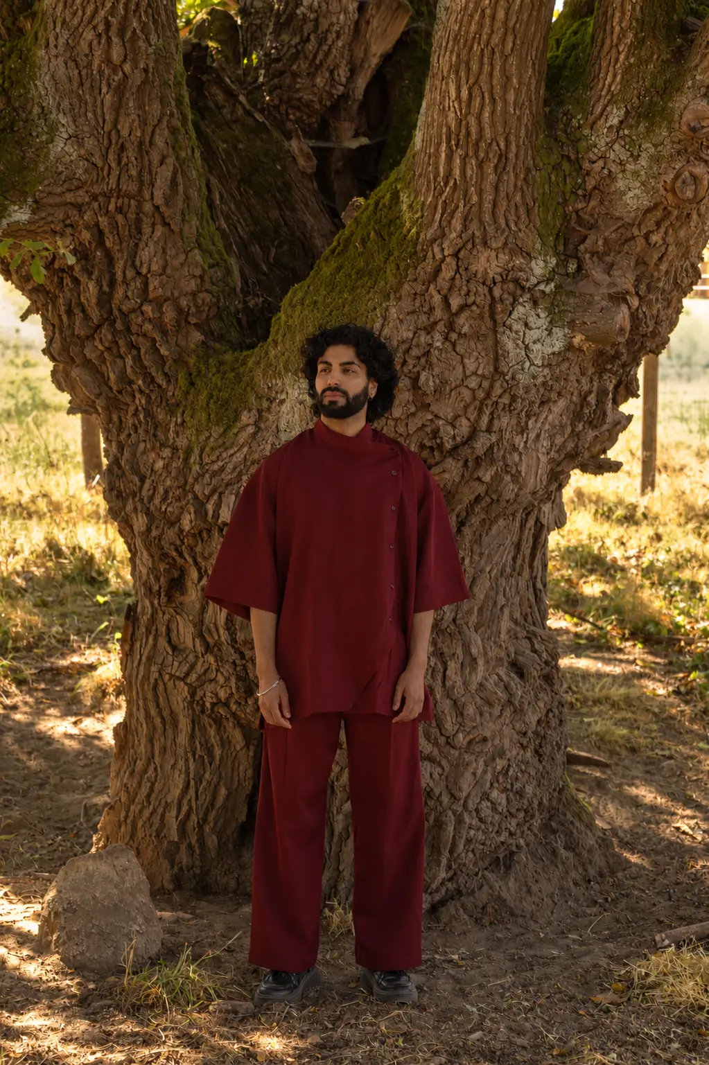 Harshit Dixit standing in a deep red costume beneath an ancient tree